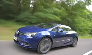 Niebieski Opel Cascada z otwartym dachem pędzi leśną drogą.