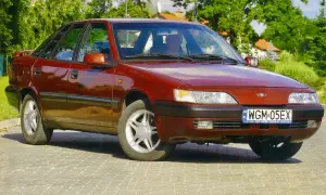 Czerwony Daewoo Espero z lat 90. stoi na brukowanej drodze, otoczony zielenią.
