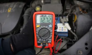 Czerwony multimetr pokazuje 12.72V przy akumulatorze samochodowym.