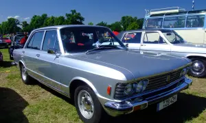 Srebrny fiat 130 z lat 70. prezentuje się elegancko na trawie, obok innych klasyków.