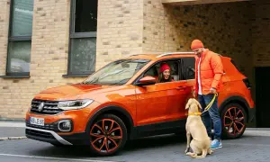 Pomarańczowy mały SUV z parą i psem. Kobieta w czerwonej czapce siedzi za kierownicą, mężczyzna w pomarańczowej kurtce trzyma psa na smyczy.
