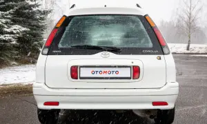 Biała Toyota Corolla E11 kombi z napisem "OTOMOTO" na tablicy rejestracyjnej, stoi na mokrym asfalcie podczas opadów śniegu.