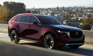 Elegancka, bordowa Mazda CX-90 sunie po drodze na tle malowniczego, miejskiego krajobrazu.