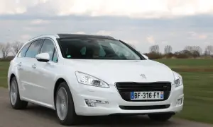Biały Peugeot 508 SW z czarnym dachem jedzie po drodze, otoczony zielonymi polami i drzewami.
