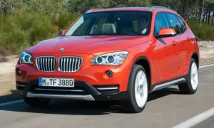 Pomarańczowy BMW X1 E84 w ruchu, z kierowcą za kierownicą, na tle drzew.