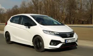 Biały Honda Fit z czarnymi felgami jedzie po drodze, otoczony drzewami.