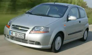 Srebrny Chevrolet Kalos w wersji 3-drzwiowej jedzie po drodze, otoczony zielenią.
