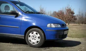 Błękitny Fiat Albea stoi na polnej drodze, w tle jesienne drzewa.
