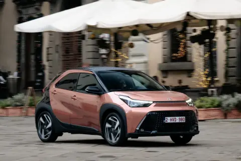 Różowy Toyota Aygo X z czarnym dachem jedzie po brukowanej ulicy.