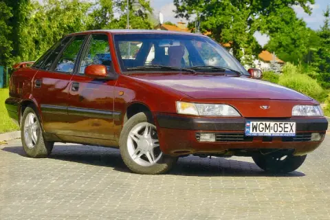Czerwony Daewoo Espero z lat 90. stoi na brukowanej drodze, otoczony zielenią.