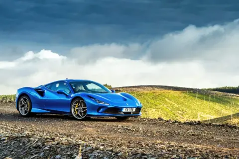 Niebieskie Ferrari F8 na tle malowniczego, pagórkowatego krajobrazu z chmurami na niebie.