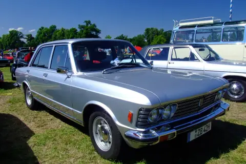 Srebrny fiat 130 z lat 70. prezentuje się elegancko na trawie, obok innych klasyków.