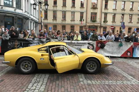 Żółte Ferrari Dino z otwartymi drzwiami, kierowca w środku, tłum widzów za barierką.