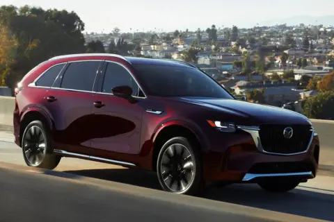 Elegancka, bordowa Mazda CX-90 sunie po drodze na tle malowniczego, miejskiego krajobrazu.