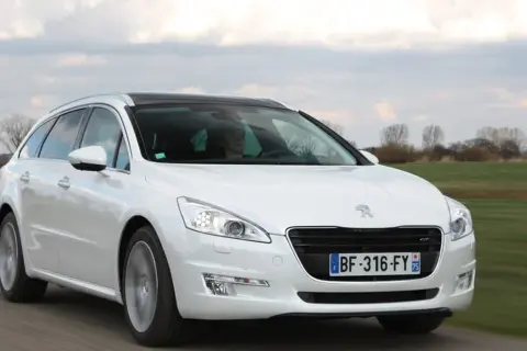 Biały Peugeot 508 SW z czarnym dachem jedzie po drodze, otoczony zielonymi polami i drzewami.