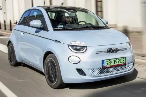 Jasnoniebieski fiat elektryczny 500e z czarnym dachem jedzie ulicą. Kierowca w okularach przeciwsłonecznych prowadzi auto.