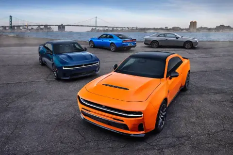 Pomarańczowy i niebieski Dodge Charger Hellcat obok szarego coupe, z mostem i miastem w tle.