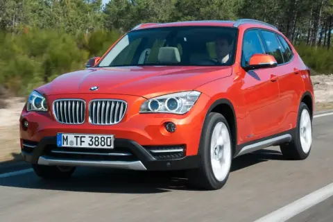 Pomarańczowy BMW X1 E84 w ruchu, z kierowcą za kierownicą, na tle drzew.