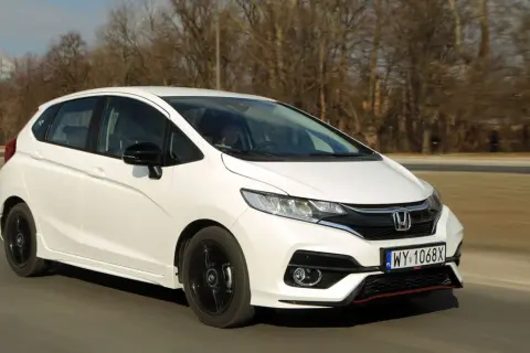 Biały Honda Fit z czarnymi felgami jedzie po drodze, otoczony drzewami.