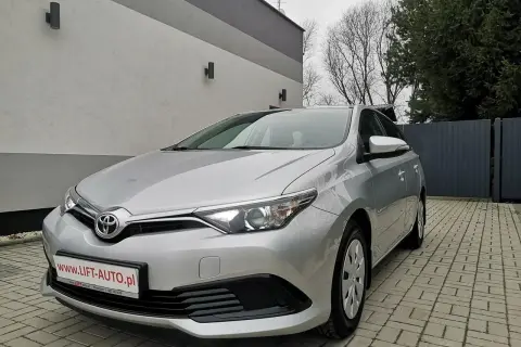 Srebrna Toyota Auris 1.6 benzyna zaparkowana na podjeździe z kostki brukowej, przed nowoczesnym budynkiem.