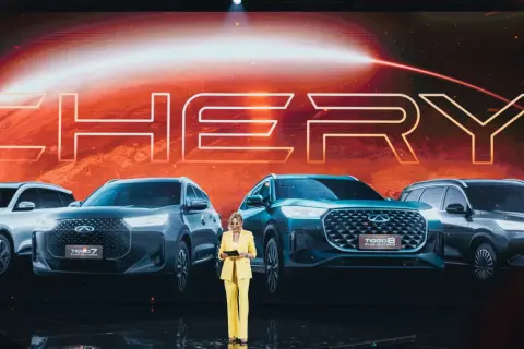 Kobieta w żółtym garniturze prezentuje nowe modele Chery Tiggo. Na ekranie za nią widnieje logo marki.
