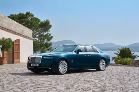 Elegancki Rolls-Royce Ghost w kolorze głębokiego turkusu zaparkowany na kamiennym dziedzińcu, z widokiem na morze i góry.
