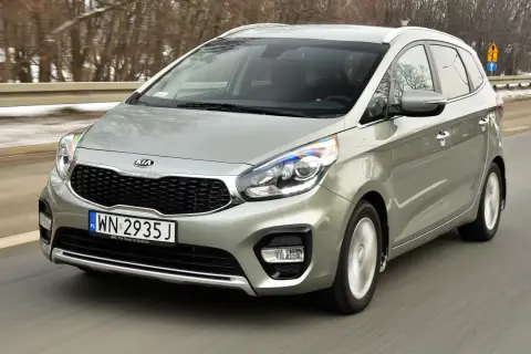 Srebrny Kia Carens 4 w ruchu drogowym, z widocznym grillem i reflektorami.