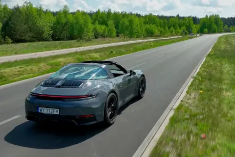 Szary Porsche Targa sunie po asfaltowej drodze, otoczony zielenią.