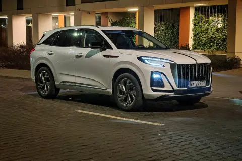 Biały Hongqi HS3 z charakterystycznym grillem i nowoczesnymi światłami stoi na mokrej, brukowanej ulicy nocą.