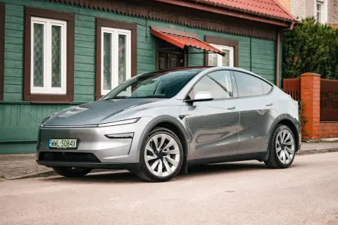 Srebrny Tesla Juniper, nowoczesny samochód elektryczny, zaparkowany przed tradycyjnym, zielonym domem z czerwonym dachem.