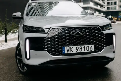 Biały Tiggo 4 z charakterystycznym grillem i pionowymi światłami LED, zaparkowany na mokrej drodze.