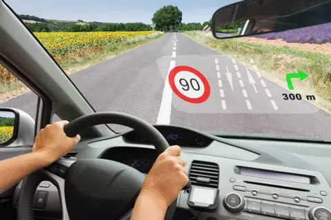 Widok z kabiny samochodu. Na szybie wyświetla się HUD z ograniczeniem prędkości 90, nawigacją i odległością 300 m do skrętu.