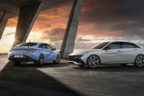 Dwa sportowe samochody Hyundai Elantra N, jeden niebieski, drugi jasnoszary, stoją pod betonowym mostem o zachodzie słońca.