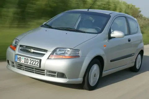 Srebrny Chevrolet Kalos w wersji 3-drzwiowej jedzie po drodze, otoczony zielenią.