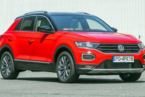 Czerwony, używany Volkswagen T-Roc z czarnym dachem i przyciemnianymi szybami, zaparkowany na tle białej ściany.