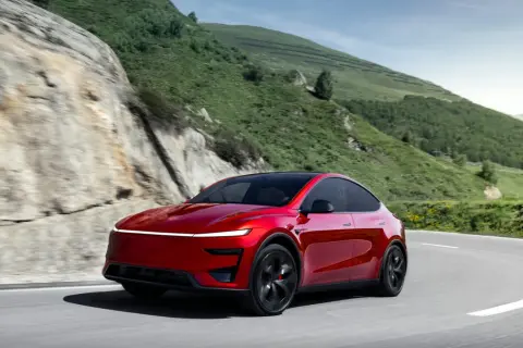 Czerwony Tesla Y Performance pokonuje zakręt na górskiej drodze, otoczony zielonymi wzgórzami i skalistymi zboczami.