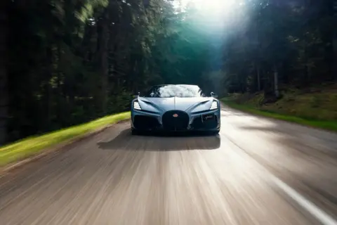 Niebieski Bugatti Mistral pędzi leśną drogą, słońce przebija się przez drzewa.