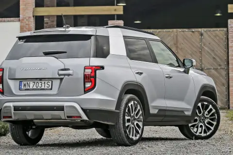 Srebrny SsangYong Torres z polskimi tablicami rejestracyjnymi WN 7031S stoi na żwirowej drodze przed drewnianą bramą.
