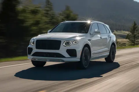 Biały Bentley SUV pędzi po krętej drodze wśród zielonych wzgórz.