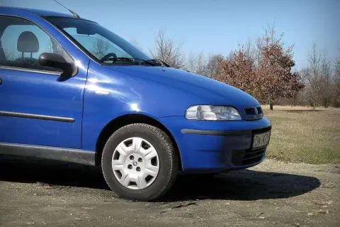 Błękitny Fiat Albea stoi na polnej drodze, w tle jesienne drzewa.