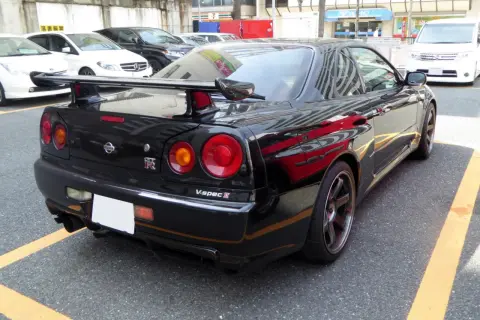 Czarny Nissan GT-R R34 V-spec II z charakterystycznymi okrągłymi światłami i dużym spojlerem, zaparkowany na ulicy.
