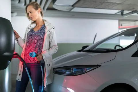 Kobieta podłącza kabel do ładowania samochodu elektrycznego w garażu podziemnym bloku.