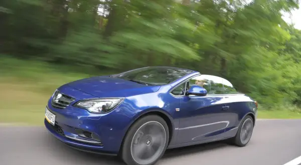 Niebieski Opel Cascada z otwartym dachem pędzi leśną drogą.
