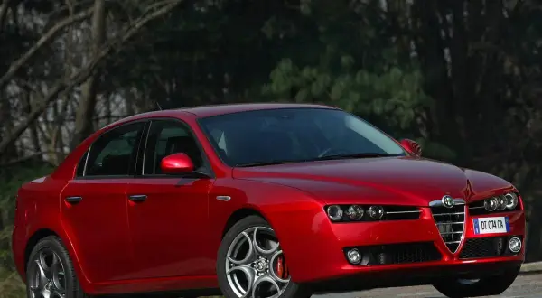 Czerwone Alfa Romeo 159 1.9 JTD z charakterystycznym grillem, zaparkowane na tle drzew.