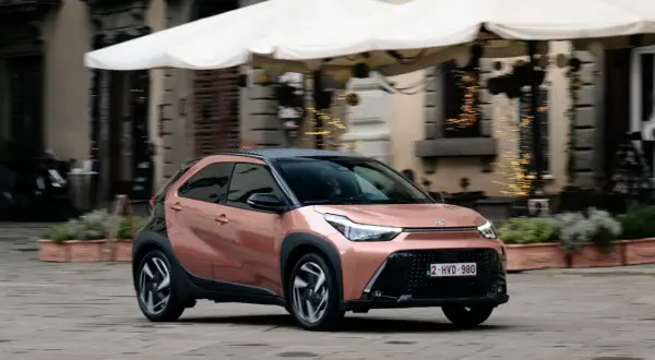 Różowy Toyota Aygo X z czarnym dachem jedzie po brukowanej ulicy.