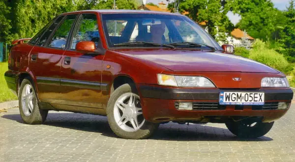 Czerwony Daewoo Espero z lat 90. stoi na brukowanej drodze, otoczony zielenią.