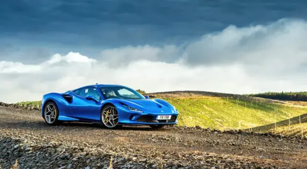 Niebieskie Ferrari F8 na tle malowniczego, pagórkowatego krajobrazu z chmurami na niebie.