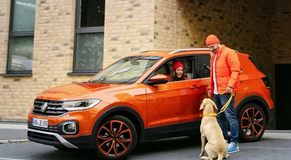 Pomarańczowy mały SUV z parą i psem. Kobieta w czerwonej czapce siedzi za kierownicą, mężczyzna w pomarańczowej kurtce trzyma psa na smyczy.