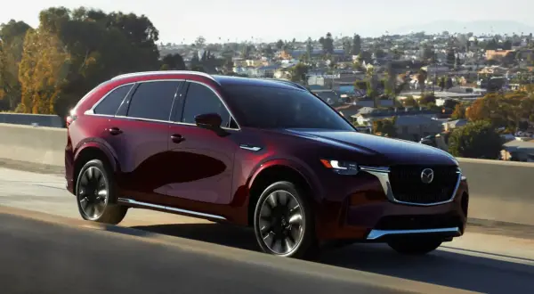 Elegancka, bordowa Mazda CX-90 sunie po drodze na tle malowniczego, miejskiego krajobrazu.
