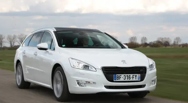 Biały Peugeot 508 SW z czarnym dachem jedzie po drodze, otoczony zielonymi polami i drzewami.